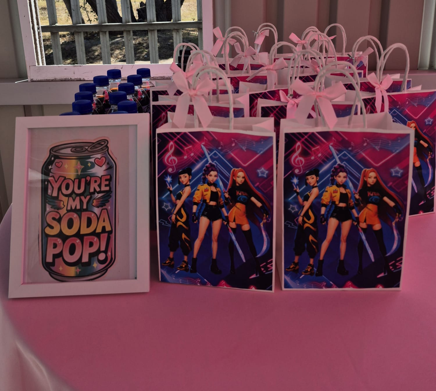 Rows of printed party favour bags with pink ribbon bows beside a framed “You’re My Soda Pop” print on a pink tablecloth