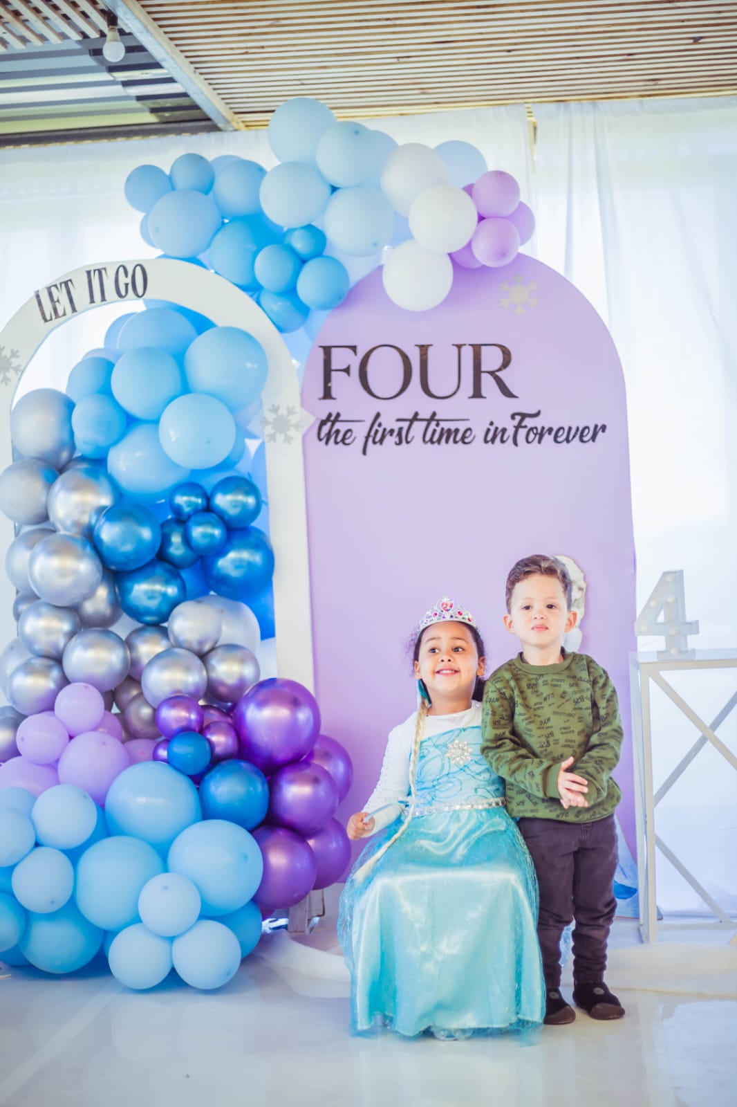 Two party guests posing in front of the lilac “FOUR – the first time in forever” backdrop