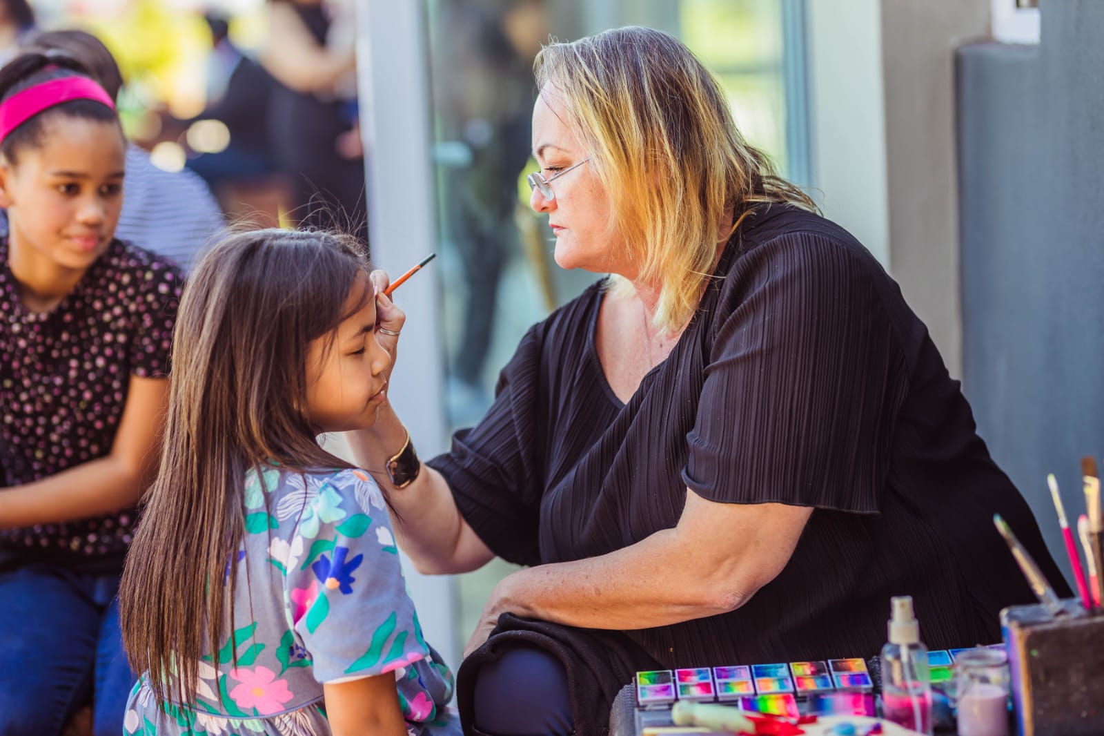 A face-painting artist decorating a young guest’s face while another child watches